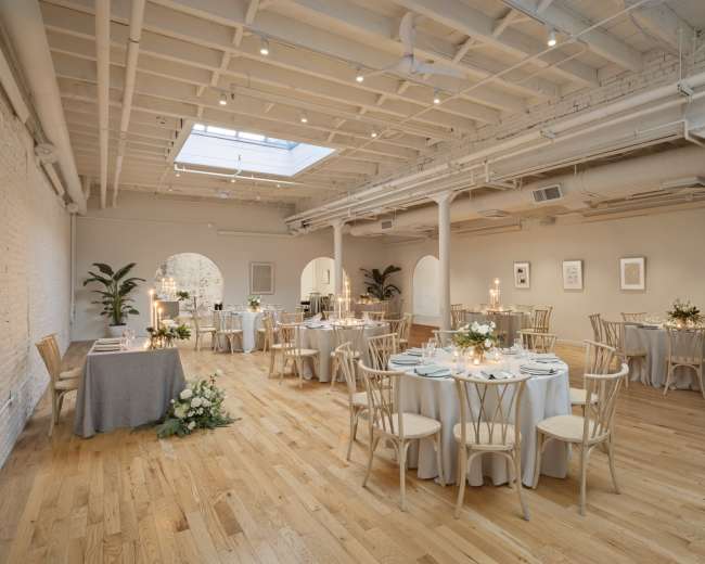 The image shows a well-lit event space with multiple circular tables set for dining, alongside elegant floral arrangements and a skylight above.