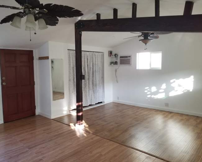 A room with wooden flooring, a mirror, a red front door, and a ceiling fan with palm leaf blades.