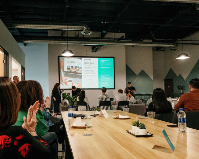 A group of people is seated at a long table in a modern conference room, facing a large screen displaying information, while some attendees applaud.