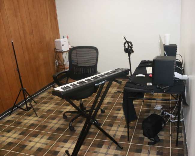 A small music recording studio features a keyboard on a folding stand, a chair, a desk with a computer, and various audio equipment.
