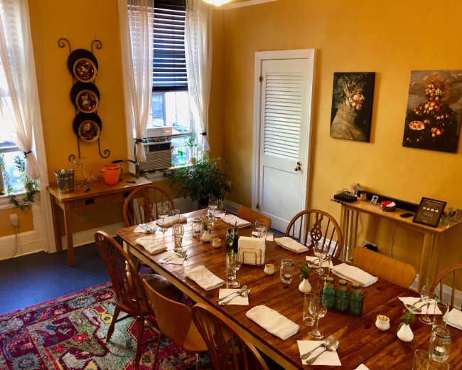 A well-set dining table is arranged with utensils and glassware in a warm, yellow-walled room featuring two pieces of framed art.