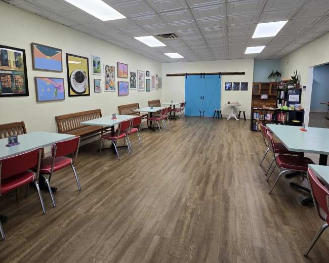 The image shows a spacious, well-lit room with numerous tables and chairs arranged neatly, along with colorful artwork on the walls and a wooden shelving unit in the background.