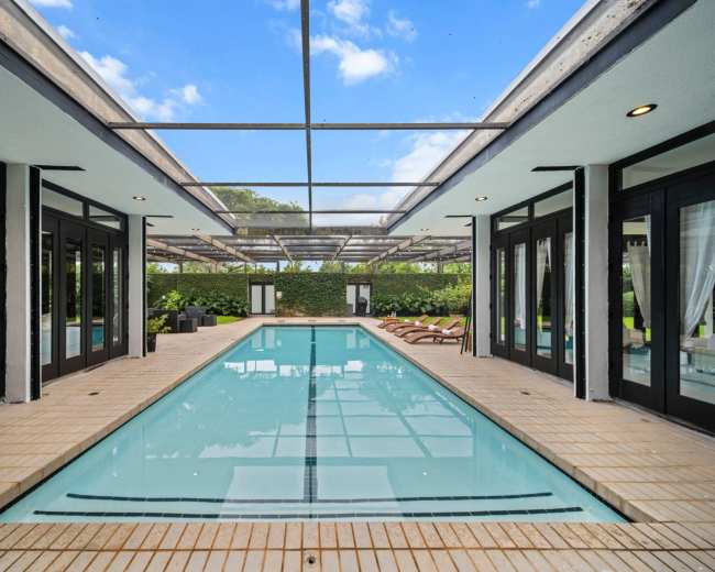 The image shows a modern indoor swimming pool surrounded by large glass doors and lounge chairs, with greenery visible outside.