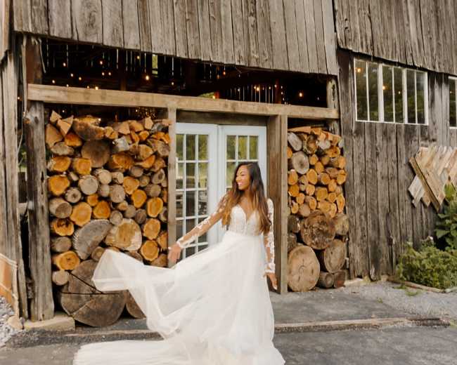 A woman in a flowing white gown poses in front of a rustic wooden barn with stacked logs.