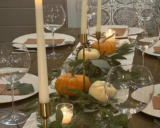 A dining table is set with glassware, candles, miniature pumpkins, and greenery as a centerpiece.