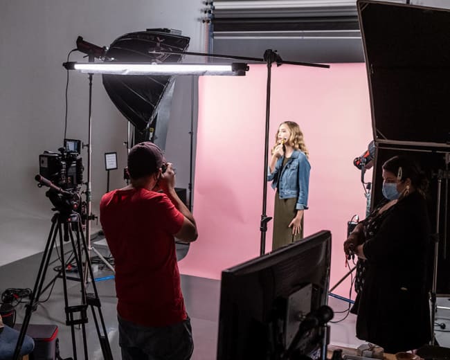 A lighting setup and filming equipment surround a model posing in front of a pink backdrop in a photography studio.