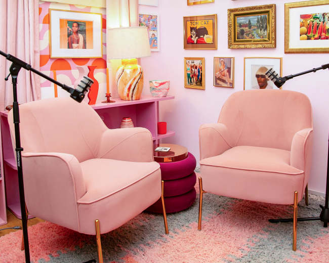 The image shows a cozy corner of a room featuring two pink armchairs facing each other, surrounded by colorful artwork on the walls and decorative items, with soft lighting provided by a lamp.