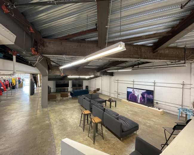 The image shows a spacious, industrial-style room with a television mounted on the wall, seating areas, and clothing racks on one side.