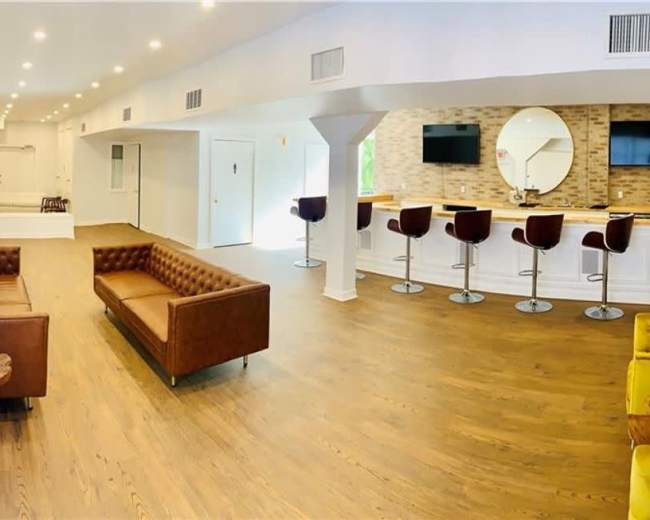 The image shows a spacious, well-lit lounge area featuring two brown leather couches, a bar with high stools, and wooden flooring.