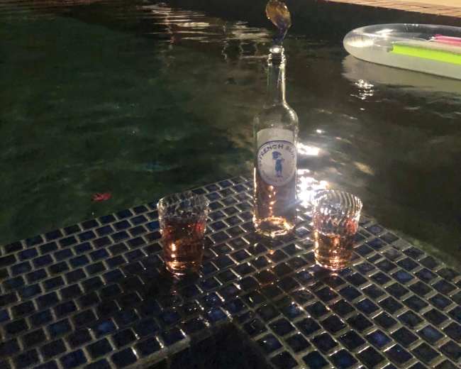 A bottle and two glasses are placed on a blue tiled surface beside a pool, with lounge chairs and a lit house in the background.