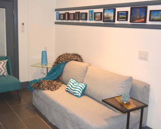 A living room with a gray sofa, a teal accent chair, a small glass side table, and a series of framed pictures mounted on the wall.