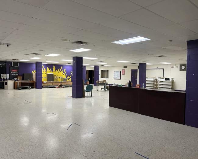 The image shows an empty cafeteria with a painted sun mural on one wall and tables stacked against the side.