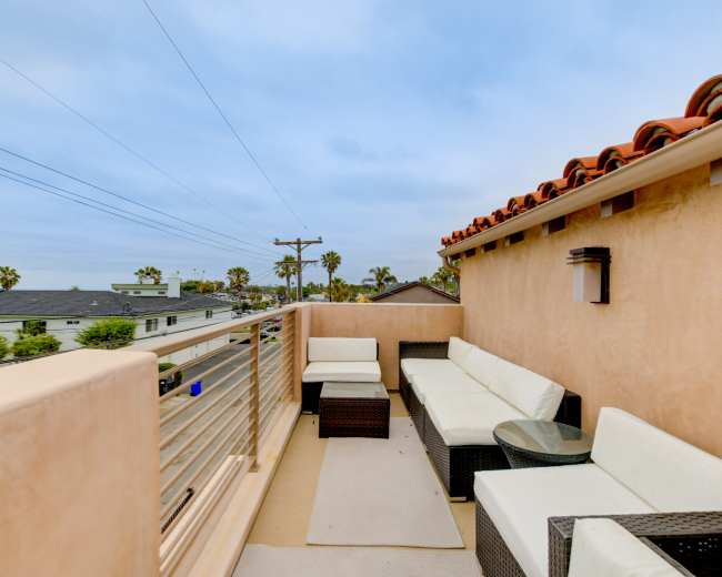 The image shows a rooftop terrace with wicker seating, a small table, and a view of nearby buildings and palm trees under a cloudy sky.