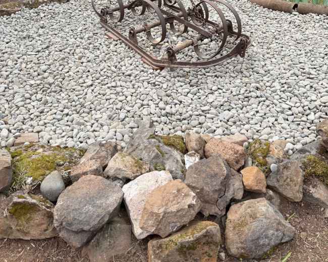 An old, rusted farming implement is displayed in a gravel-covered garden surrounded by stones and greenery.
