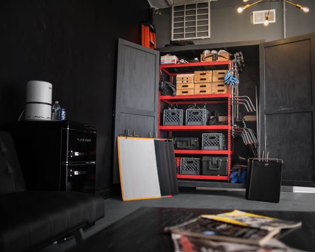 A storage area features a red shelving unit filled with boxes and equipment, alongside a black couch and a small fridge.