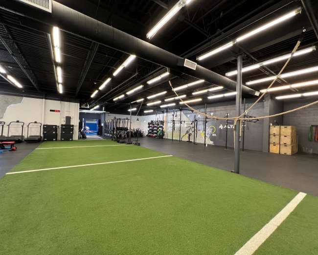 The image shows a spacious indoor gym with artificial turf, workout equipment, and storage boxes.