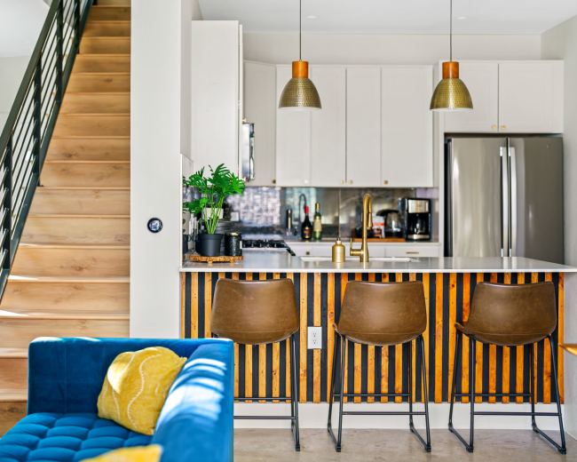 The image shows a modern kitchen area with a stairway to the left, featuring a bar with three stools and a countertop adorned with plants and kitchen appliances.