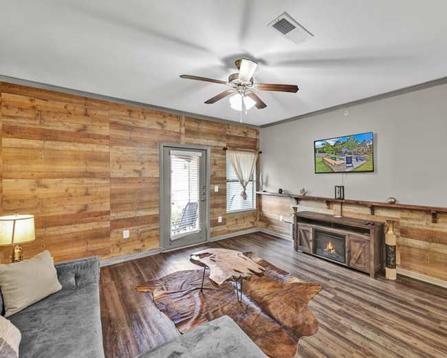 The living room features wooden walls, a ceiling fan, a TV mounted above a decorative fireplace, and a large area rug.