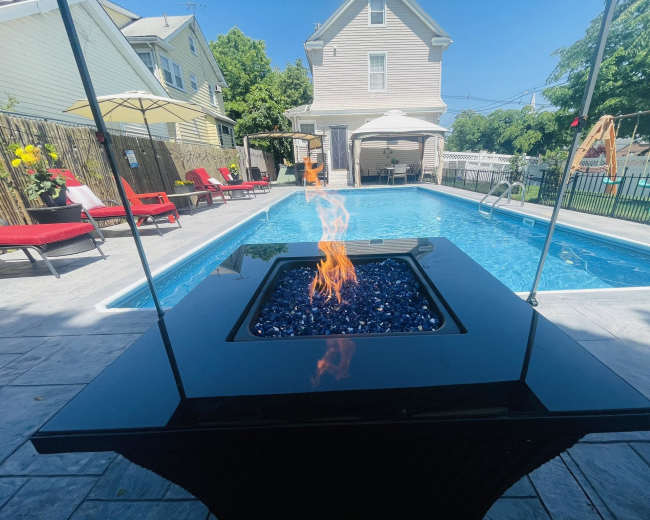 A modern black fire table with vibrant blue glass sits in front of a clear swimming pool surrounded by red lounge chairs and a wooden fence.