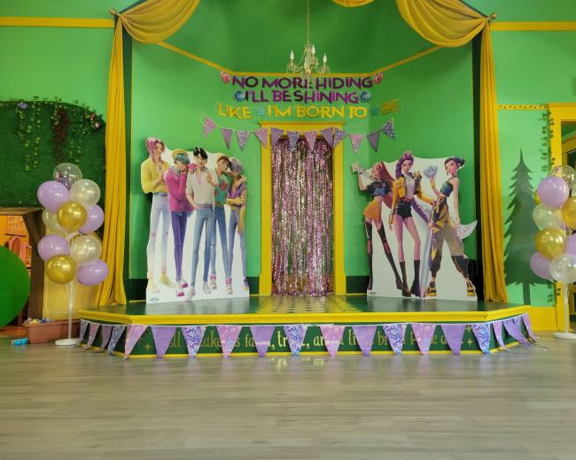 The image shows a brightly decorated stage with green walls, yellow drapery, and vibrant banners, featuring lifelike cutouts of characters and colorful balloons.
