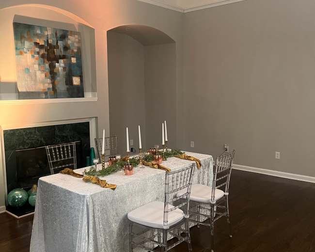 A dining table covered with a silver tablecloth is set with candles and decorations in a well-lit room featuring a fireplace and large windows.