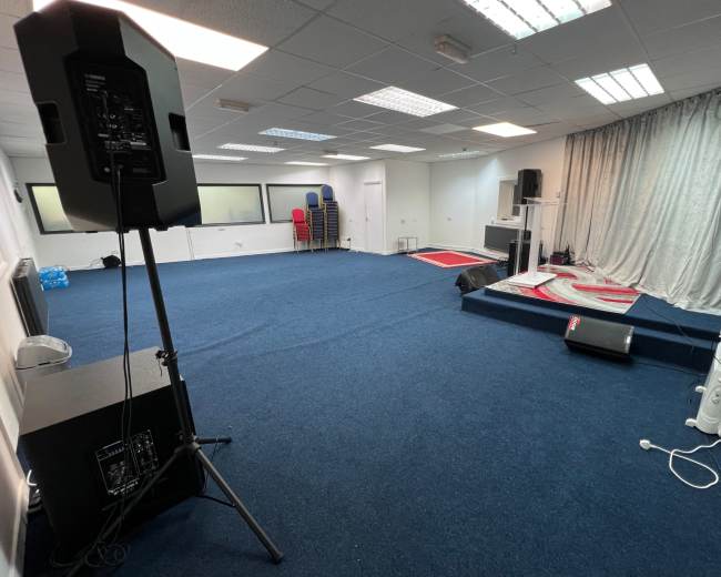The image shows a spacious room with blue carpet, featuring a stage with a podium and curtains, audio equipment on either side, and a few chairs stacked in the background.