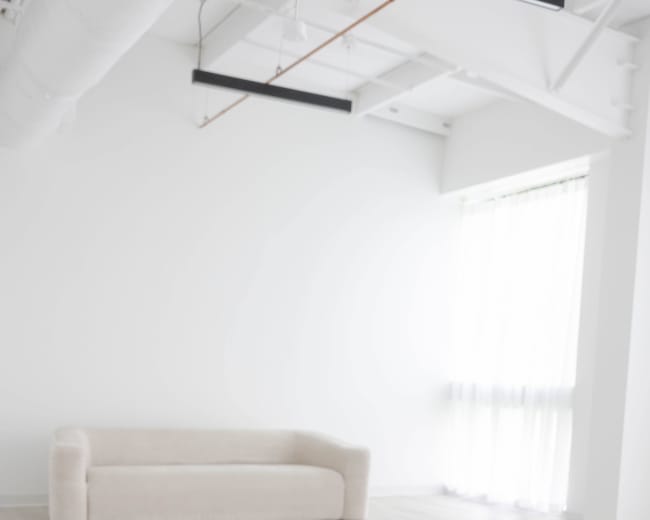 A light-filled, minimalist room features a beige sofa against a white wall, with large windows allowing natural light to stream in.