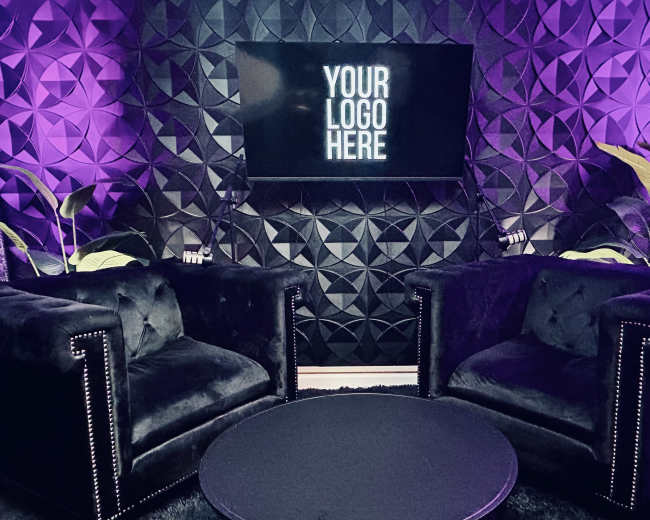The image shows a modern seating area with two black velvet armchairs and a round table, set against a textured wall featuring purple lighting and a television displaying the text "YOUR LOGO HERE."