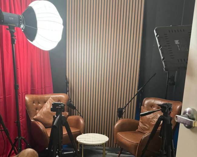 The image shows two brown leather chairs positioned around a small white table, facing a backdrop of vertical wooden panels, illuminated by a softbox light and equipped with camera tripods and microphones.
