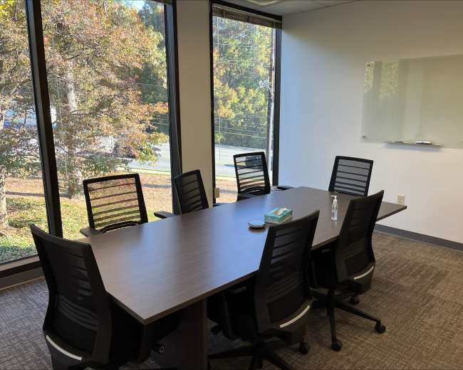 A modern conference room features a large table surrounded by six ergonomic chairs, with large windows showing a view of trees outside.