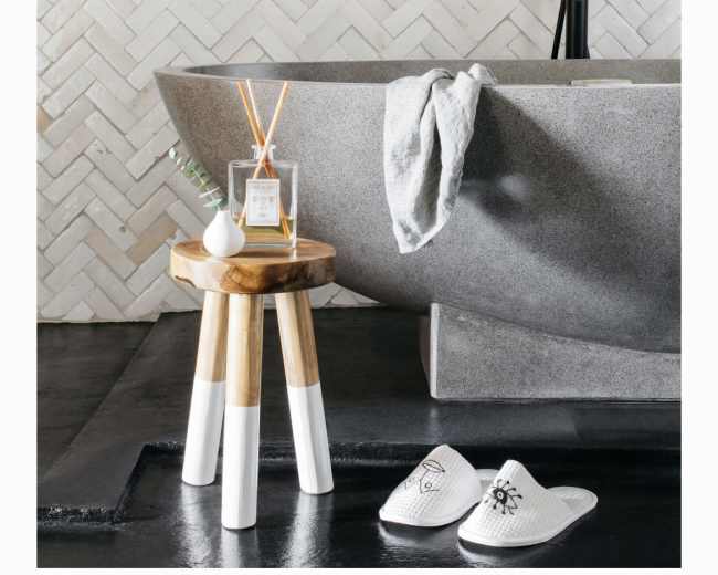 A modern bathroom features a stone bathtub beside a wooden stool with a scented candle and reed diffuser, set against a herringbone tile wall.