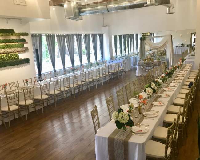 A banquet hall decorated with tables set for a formal event, featuring white tablecloths, floral centerpieces, and large windows with sheer curtains.