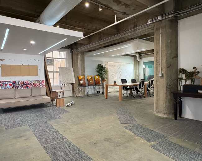 The image shows a spacious office area with a sofa, a large table surrounded by chairs, and artworks displayed on easels against a concrete wall.