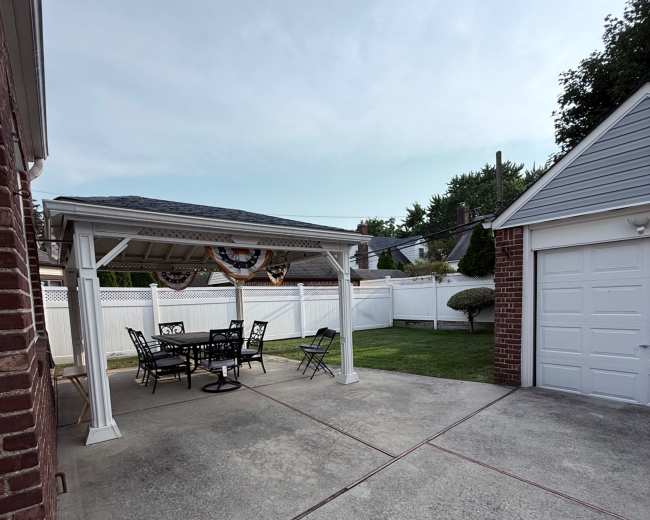 The image shows a covered patio with a black metal table and chairs, adjacent to a brick garage and enclosed by a white fence.