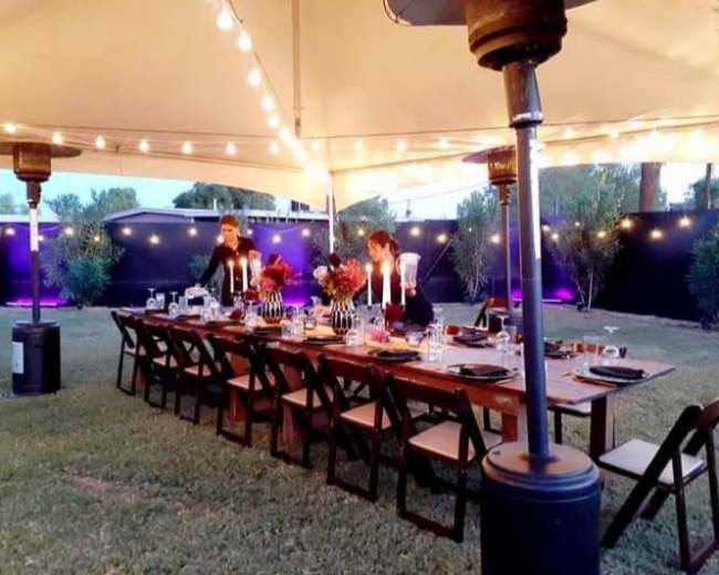 A long wooden dining table is set under a large tent, decorated with candles and flowers, surrounded by chairs and illuminated by string lights.