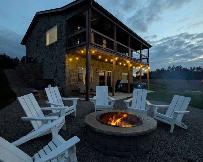 A stone house with a multi-level design features a fire pit surrounded by several white Adirondack chairs, illuminated by string lights in the evening sky.