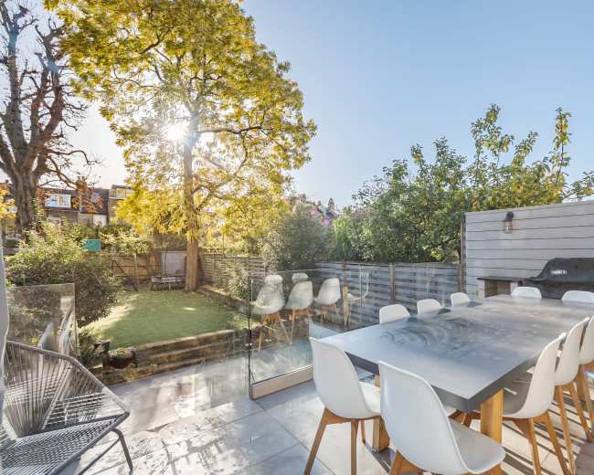 A modern outdoor dining area features a large table surrounded by white chairs, with a lush garden and towering trees in the background.