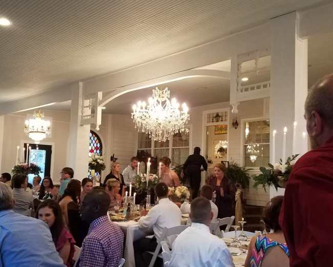 The scene depicts a dining event in a well-decorated interior with guests seated at tables and a chandelier hanging above.