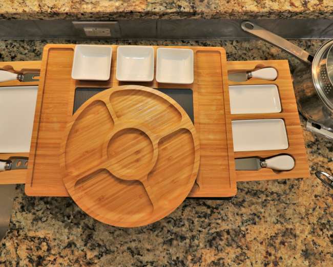 A bamboo serving platter with multiple small dish compartments is arranged on a granite countertop, accompanied by utensils.