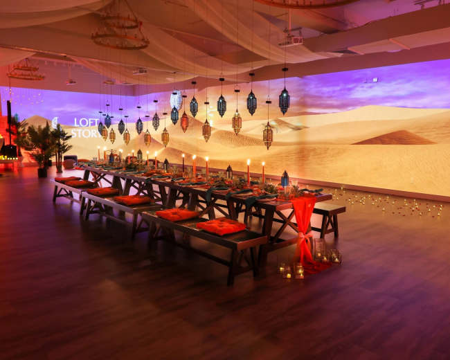 The image shows a beautifully arranged dining setup with a long table adorned with decorations and surrounded by lanterns, set in a space featuring a desert-themed projected backdrop.