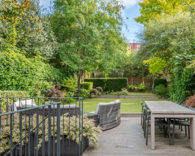 A well-maintained garden features a dining table and chairs on a deck, surrounded by lush greenery and neatly trimmed hedges.