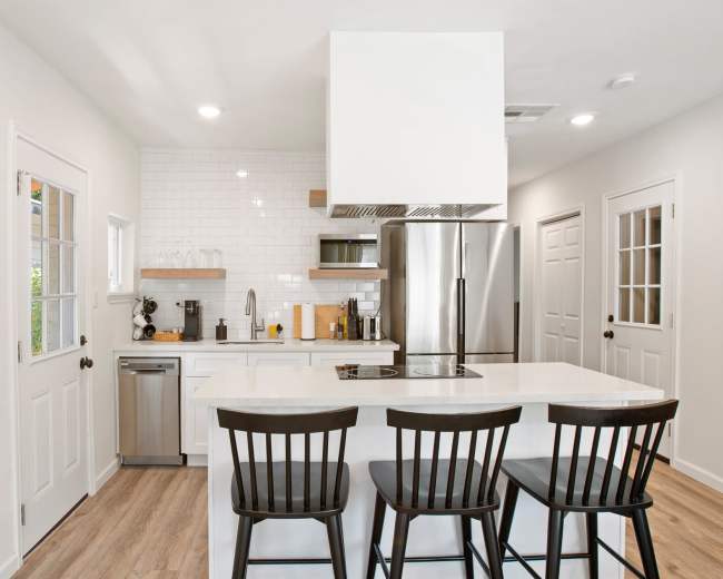 The image shows a modern kitchen with white cabinetry, a stainless steel refrigerator, and three black bar stools at a white countertop.
