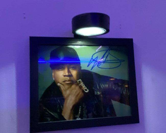 The image shows a framed, autographed portrait of a man in a black hat and jacket, illuminated by a mounted light on a purple wall.
