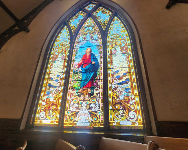 The image shows a brightly colored stained glass window depicting a figure in a red and blue robe, set within a church interior.