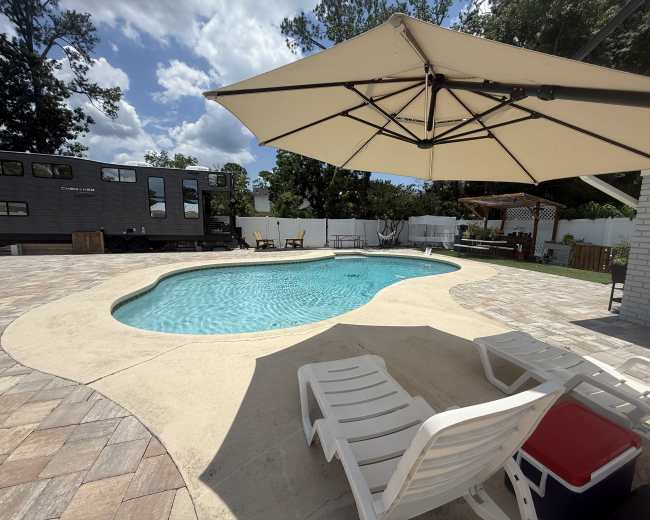 The image features a swimming pool surrounded by a patio with lounge chairs, an umbrella, and an RV parked nearby.