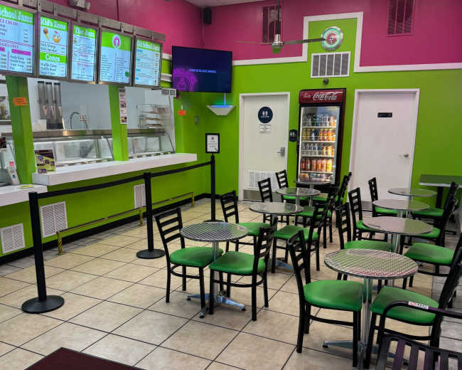 The image shows a brightly colored fast-casual restaurant with green and pink walls, featuring several tables and chairs arranged in the dining area.