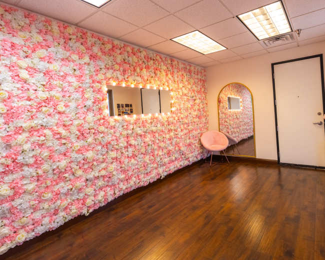 The image shows a room with a floral wall made of pink and white flowers, a mirror, and a chair, all set against a wooden floor.