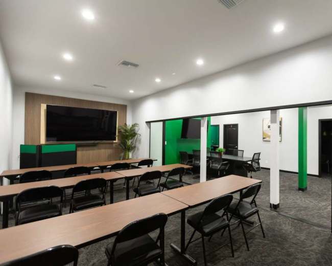 The image shows a modern meeting room with a series of long tables and chairs arranged facing a large screen and wall-mounted television.