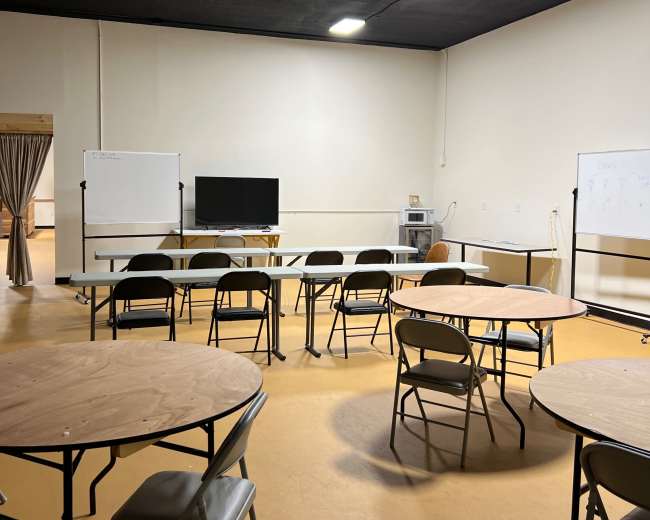 The image shows a spacious room set up with circular tables and folding chairs arranged around a central area, featuring a whiteboard and a television screen.