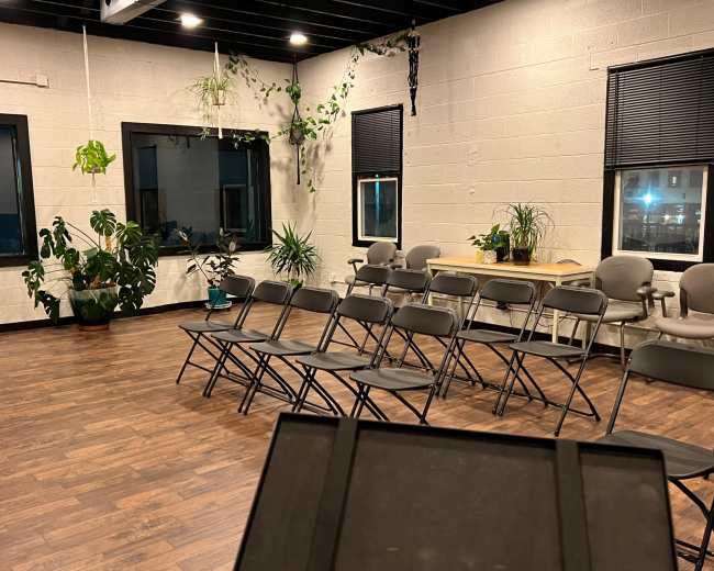 A spacious room with folding chairs arranged in rows, large windows, plants, and a table along one wall.